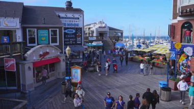 İnsanlar Balıkçı İskelesi, Monterey 'deki canlı sahil yolunda yürüyorlar. Dükkanları, renkli tabelaları ve arka planda botlarla dolu bir marinası var..