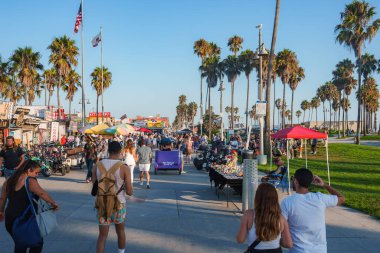 Venice Beach sahil şeridi insanlarla, satıcılarla ve palmiyelerle dolup taşıyor. Amerikan ve Kaliforniya bayrakları açık mavi gökyüzünün altında renkli şemsiyelerle dalgalanıyor..