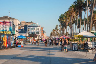 İnsanlar Los Angeles 'taki canlı Venice Beach Boardwalk boyunca yürüyüp bisiklet sürüyor. Palmiye ağaçları yol boyunca uzanır, duvarları renkli grafiti sanatıyla kaplıdır..