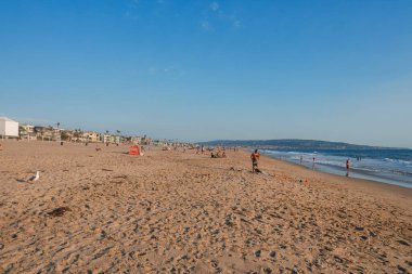 İnsanlar Venedik Sahili 'nin Los Angeles' taki kumlu kıyı şeridinde güneşin tadını çıkarıyorlar. Yerleşim binaları ve palmiye ağaçları açık mavi gökyüzünün altında kıyı boyunca uzanır..