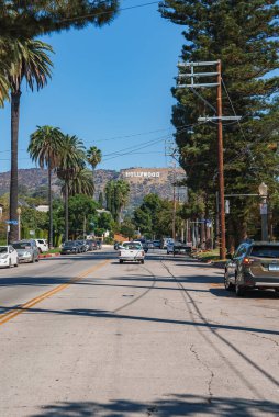 Hollywood tabelası arka plandaki tepelerde duruyor. Önünde palmiye ağaçları ve arabalarla dolu bir Los Angeles sokağı açık mavi gökyüzünün altında..