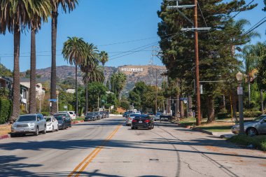 Los Angeles caddesi palmiye ağaçları ve park halindeki arabalarla doludur. Güneşli bir günde tepelerde mavi gökyüzünün altında Hollywood tabelası görünür..