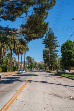 Los Angeles caddesinde uzun palmiye ağaçları ve park edilmiş arabalar var. Tepelerde mavi gökyüzünün altında ikonik Hollywood işareti var..