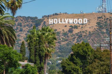 İkonik Hollywood tabelası Los Angeles tepelerinde duruyor. Kuru arazi ve yemyeşil alanlarla çevrili ve elektrik direkleri yakınlardaki konutları işaret ediyor..