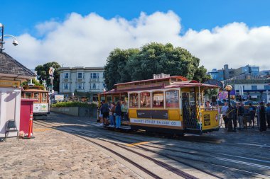 Çevresi insanlarla çevrili klasik bir San Francisco tramvayı. Parke taşı sokaklar ve şehir binaları berrak mavi bir gökyüzünün altında görülebilir..