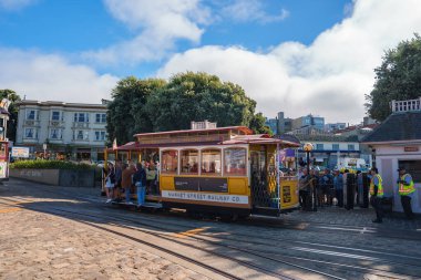 Klasik bir San Francisco teleferiği, parke taşlı bir caddede insanlarla çevrili bir pikapta konumlandırılmıştır. Ağaçlar ve çok katlı binalar parçalı bulutlu bir gökyüzünün altında görülebilir..