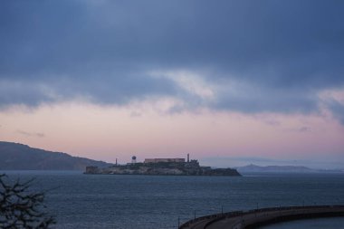 Alcatraz Adası, mavi ve pembe renkleriyle sakin bir alacakaranlık gökyüzüne karşı gölgelenmiştir. Sakin sular ve uzak tepelerle çevrili tarihi hapishane binaları görülebilir..