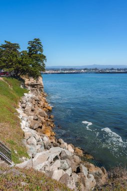 Çimenli alanları ve ağaçları olan kayalık bir kıyı şeridi okyanusa uzanan uzun bir iskeleye yol açar. Şehir manzarası ve uzak dağlar berrak bir gökyüzünün altında görülebilir..