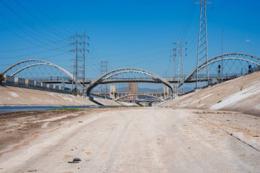 The iconic Los Angeles River features a concrete channel, arched bridges, and towering electrical pylons under a clear blue sky, highlighting the urban landscape. clipart