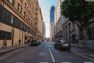 Los Angeles şehir merkezinde hareketli bir caddede yüksek binalar, mavi camdan bir gökdelen, park edilmiş arabalar, yayalar, yayalar ve bir ağaç bulunuyor..