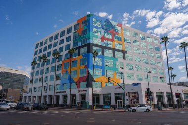 Renkli bir geometrik duvar resmi Los Angeles 'taki modern bir binayı süslüyor. Sokak boyunca uzanan uzun palmiye ağaçları ve zemin katta görünen bir restoran..