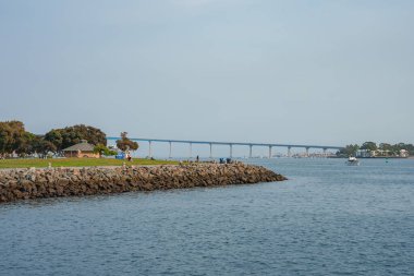 Coronado Köprüsü, San Diego, Kaliforniya 'da su boyunca uzanır. Ağaçlı kayalık bir kıyı şeridi ve ön planda bir bina, suda bir tekne var..
