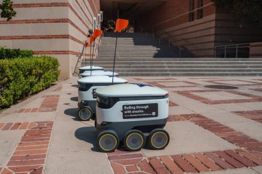 Turuncu bayraklı bir grup teslimat robotu UCLA 'da bir tuğla kaldırım hattında. Basamaklar ve çitler sahne çerçevesi, bir üniversite ortamında sergilenen teknoloji.