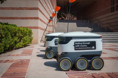 Bayraklı iki otonom dağıtım robotu UCLA 'da asfalt kaldırımda. Sahnede tuğla ve taş mimari ve bir binaya giden basamaklar yer alıyor..