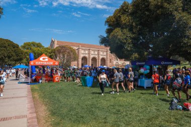 A large crowd gathers on UCLA's grassy area for a festive event. The scene includes a Gatorade tent, balloons, and the university's brick facade building. clipart