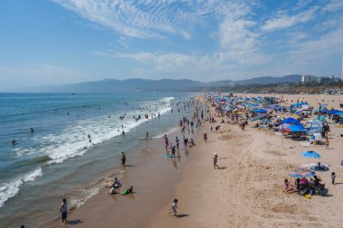 Los Angeles 'taki Venice Beach güneşlenenlerle, şemsiyelerle ve plaj havlularıyla dolu. Yüzücüler ve sörfçüler okyanusun tadını çıkarırlar, Santa Monica Dağları görünür..