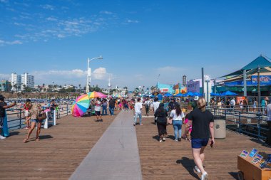 Kalabalık, Santa Monica İskelesi, Los Angeles 'ta Pasifik Park' ın dönme dolabı ve lunapark trenleriyle güneşli bir günün tadını çıkarıyor. Okyanus ve açık gökyüzü sahneyi tamamlıyor..