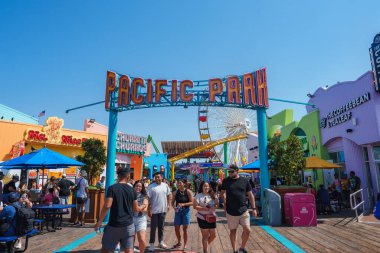 Ziyaretçiler, Santa Monica İskelesi 'ndeki canlı Pasifik Parkı tabelasının altında yürüyorlar. Dönme dolap ve lunapark treni görülebiliyor..