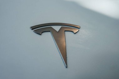 Görüntü merkezi bir Tesla logosunu gösteriyor, pürüzsüz metalik bir yüzeyde stilize edilmiş bir T amblemi. Açık gri arka plan logonun parlak tasarımını geliştirir.