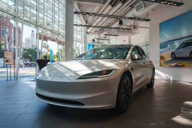 Santa Monica 'daki bir galeride parlak beyaz bir elektrikli araba sergileniyor. Büyük cam pencereler dışarıdaki palmiye ağaçlarını gösterirken, bir plaj manzarası duvarı süslüyor..