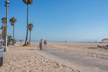 Bir bisikletçi Venedik Plajı, Los Angeles 'ta palmiye ağaçlarıyla çevrili bir patika boyunca gidiyor. Sahile gidenler voleybol ağları ve renkli bir şemsiyeyle kumun tadını çıkarıyorlar..