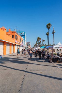 İnsanlar Venice Sahili 'ndeki renkli mağazaların ve tezgahların önünden geçiyor. Uzun palmiye ağaçları açık mavi gökyüzünün altında gölgeler bırakır..