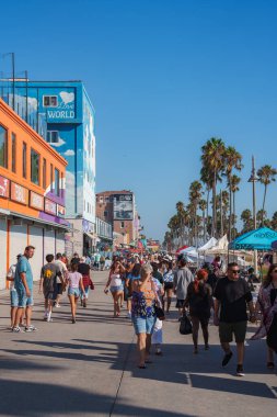 Venice Beach sahil şeridi insanlarla, renkli mağazalarla ve palmiye ağaçlarıyla dolup taşıyor. Bir duvar resminde açık mavi gökyüzünün altında 