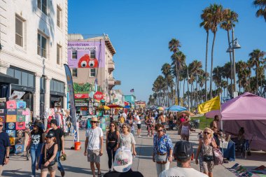 People enjoy a sunny day at Venice Beach, Los Angeles, with street vendors, colorful tents, and tall palm trees. A mural adds artistic flair to the scene. clipart