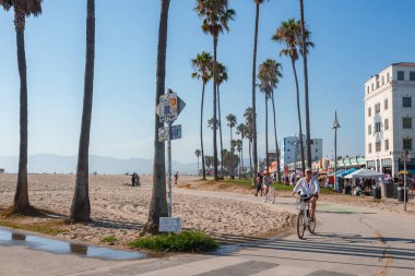Los Angeles 'taki Venedik Plajı' nda uzun palmiye ağaçları bisikletçiler ve yayalar, renkli binalar ve uzak dağları olan açık mavi bir gökyüzü bulunur..