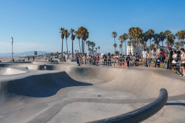 Los Angeles 'taki Venice Beach Paten Parkı' nda patenciler numaralar yapıyor. Arka planda palmiye ağaçları ve okyanusla birlikte izleyiciler çevreyi gözlüyor..