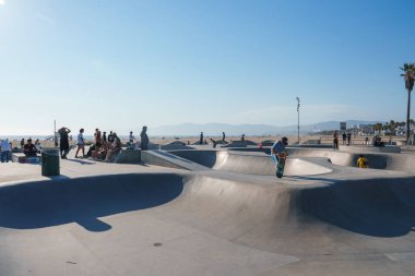 Kaykaycılar Venice Beach Paten Parkı 'ndaki rampalarda numaralar yaparlar. Okyanus ve Santa Monica Dağları manzarayı güzelleştiren bir palmiye ağacıyla görülebilir..