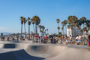 İnsanlar Venice Beach, Los Angeles 'taki bir kaykay parkında toplanıp patencilerin numaralarını izliyorlar. Uzun palmiye ağaçları ve binalar güneşli arka planda..