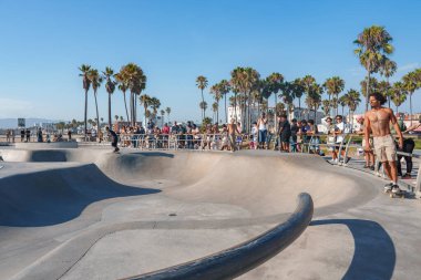 Kaykaycılar Venice Beach Kaykay Parkı 'ndaki düz beton kaselerde geziniyorlar. İzleyiciler uzun palmiye ağaçlarının altında güneşli Güney Kaliforniya gününün tadını çıkarıyorlar..