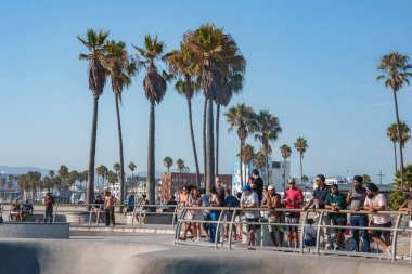 İnsanlar Venice Beach, Los Angeles 'ta bir tırabzanda toplanıp parktaki patencileri izliyorlar. Uzun palmiye ağaçları ve açık mavi bir gökyüzü güneşli günü aydınlatır..
