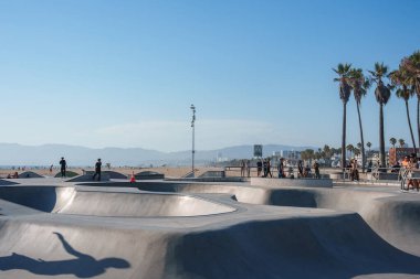 Venice Beach kaykay parkında düz beton rampalar ve kaseler var. Patenciler ve seyirciler, palmiye ağaçları, kumlu plajlar ve uzak dağlarla sahnenin tadını çıkarıyorlar..