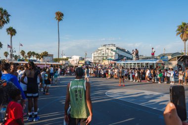 Venice Beach, Los Angeles 'ta canlı bir sahne, açık hava sahasında bir basketbol maçı. Arka planda uzun palmiye ağaçları ve hareketli tahta kaldırım görülebilir..