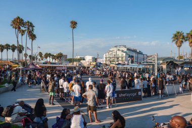 Venedik Sahili, Los Angeles 'ta çeşitli kalabalıklar toplanıyor. Palmiye ağaçları, satıcı çadırları ve açık mavi gökyüzü altında modern sahil binaları bulunuyor..