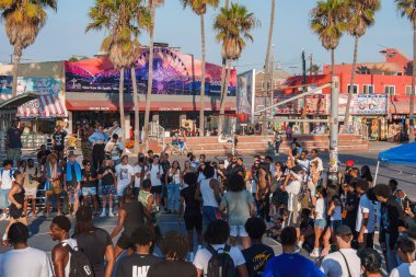Venice Beach, Los Angeles 'ta çeşitli bir kalabalık basketbol sahasının etrafında toplanıyor. Renkli duvar resimleri, palmiye ağaçları ve dönme dolap billboardları canlı görünmesini sağlar..