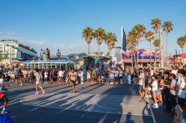 Venice Beach, Los Angeles 'ta canlı bir basketbol maçı var. Oyuncular büyük bir kalabalık, simgesel palmiye ağaçları ve arka planda açık mavi bir gökyüzü..