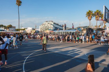 Venice Beach, Los Angeles 'ta canlı bir sahne. Bir basketbol sahasının, palmiye ağaçlarının ve açık mavi gökyüzünün altında eşsiz bir mimarinin etrafında kalabalık var..