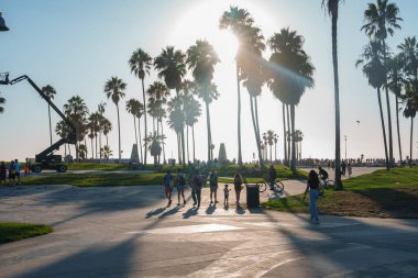 Los Angeles 'taki Venedik Plajı' nda palmiye ağaçlarının arkasında gölgeler bırakan bir günbatımı vardır. Bisikletçiler ve yayalar arka planda bir vinçle gezintinin tadını çıkarıyorlar..