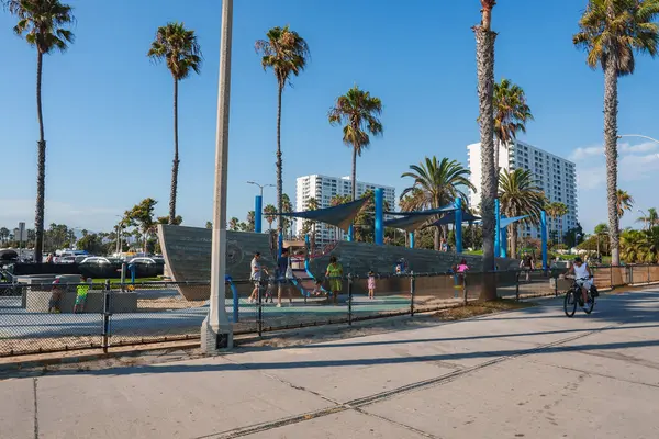 Çocuklar Venedik Plajı, Los Angeles 'ta gemi temalı bir oyun alanında oynuyorlar. Uzun palmiye ağaçları ve modern yüksek binalar canlı sahneyi çerçeveler..