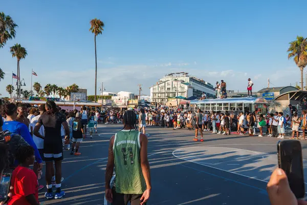 Venice Beach, Los Angeles 'ta canlı bir sahne, açık hava sahasında bir basketbol maçı. Arka planda uzun palmiye ağaçları ve hareketli tahta kaldırım görülebilir..