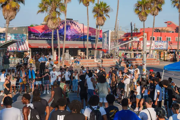 Venice Beach, Los Angeles 'ta çeşitli bir kalabalık basketbol sahasının etrafında toplanıyor. Renkli duvar resimleri, palmiye ağaçları ve dönme dolap billboardları canlı görünmesini sağlar..