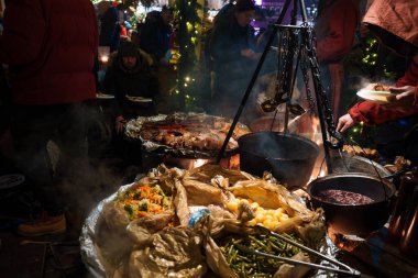 Riga 'nın Noel pazarındaki hareketli bir gıda standında renkli tabakları olan büyük tavalar yer alıyor. Şenlikli ışıklar, insanların canlı atmosferden zevk aldığı sahneyi süslüyor..