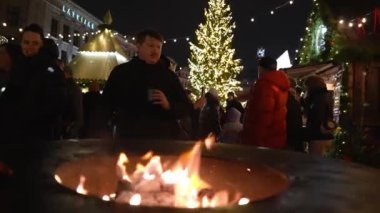 Ziyaretçiler Riga Noel pazarındaki bir şöminenin etrafında toplanıyor. Arka planda süslü tezgahlar ve Letonya binaları bulunuyor..