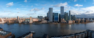 Canary Wharf 'ın modern gökdelenleri, One Canada Meydanı da dahil, Thames Nehri ön planda parçalı bulutlu bir gökyüzünün altında..