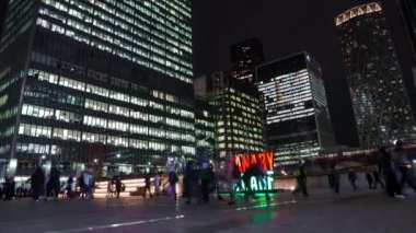 İnsanlar meydanda yürürken Canary Wharf 'ın gökdelenleri gökyüzünü aydınlatıyor. Kırmızı ve yeşil bir tabela ve çift katlı bir otobüs şehir manzarasını güçlendiriyor.