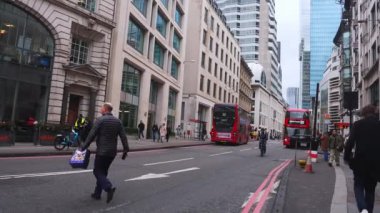 Kırmızı çift katlı otobüsler, yayalar ve bulutlu gökyüzü altında modern ve klasik binaların karışımı ile dolu bir Londra sokak sahnesi..
