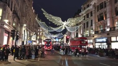 Londra 'nın Regent Caddesi' nde, tarihi mimariyi modern dekorla harmanlayan, hareketli bir tatil kalabalığı arasında melek ışığı ve ikonik kırmızı otobüsler yer alıyor..